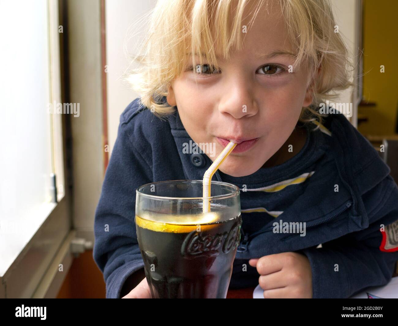 Boy 6-8 years infant child blond happy impish cheeky drinking through a straw a branded glass of Coca Cola 'COKE' with a slice of lemon added. Stock Photo