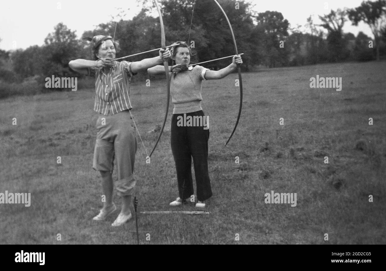 1950s, historical, outside in a field, two women, one wearing a pair of slacks, the other with the bottom of her trousers rolled up, standing with their bows and arrows doing archery. Both a sport and a fun leisure activity, in archery one uses a bow to try and to shoot arrows as close to the centre of a target as possible. Stock Photo