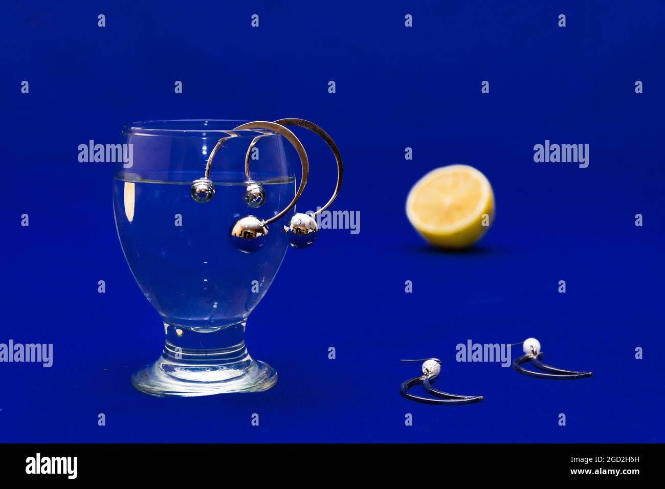 Pairs of silver earrings, a glass of water, and a slice of lemon on a blue surface Stock Photo