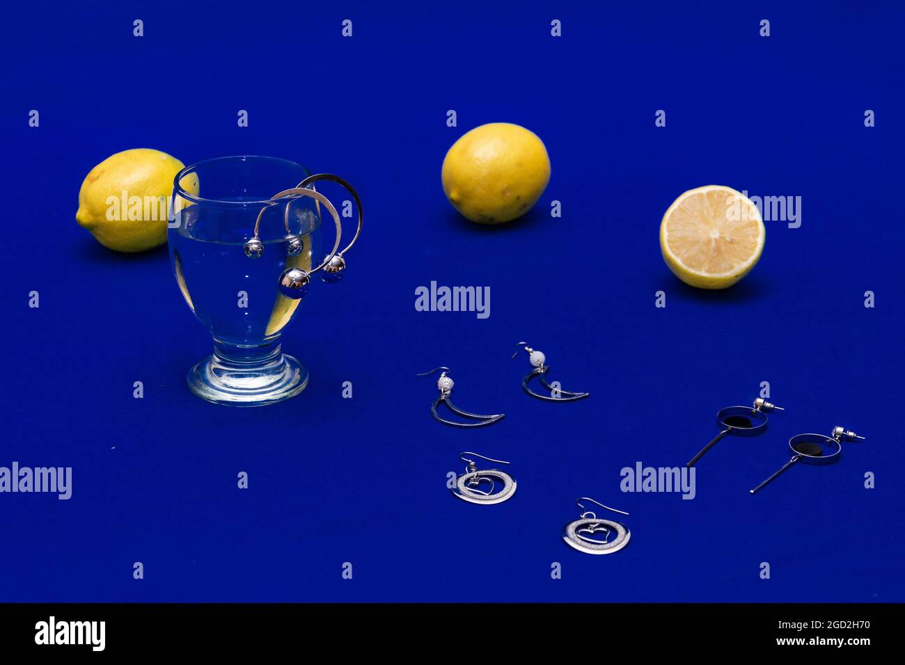 Pairs of silver earrings, a glass of water, and slices of lemon on a blue surface Stock Photo