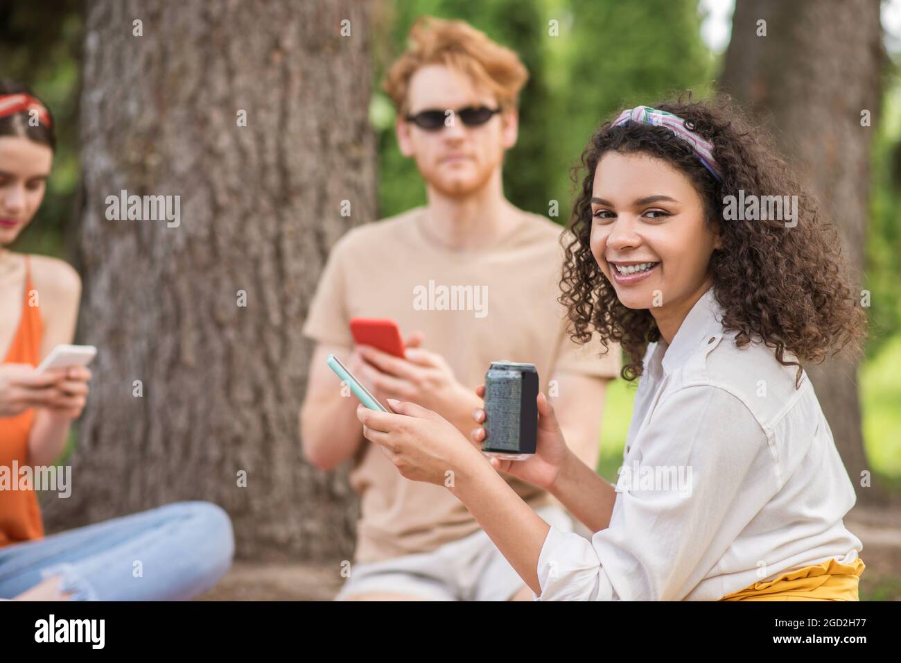 Mulatta girl with drink and smartphone and friends behind Stock Photo