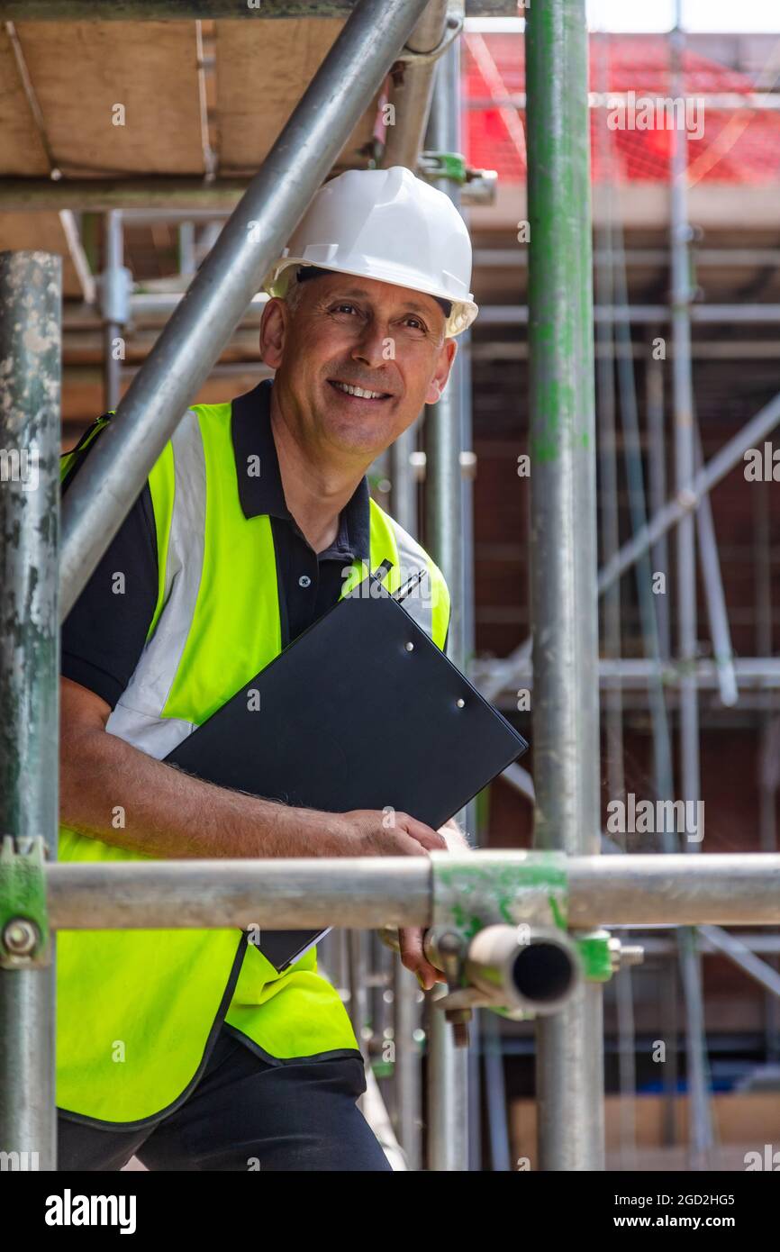 Male builder foreman, smiling happy construction worker or site manager holding a clipboard, wearing a white hard hat and hi vis vest Stock Photo