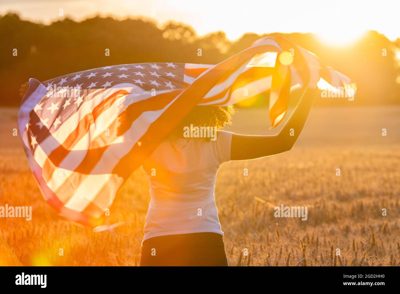 Mixed race African American girl teenager female young woman in a field of wheat or barley crops wrapped in USA stars and stripes flag in golden sunse Stock Photo