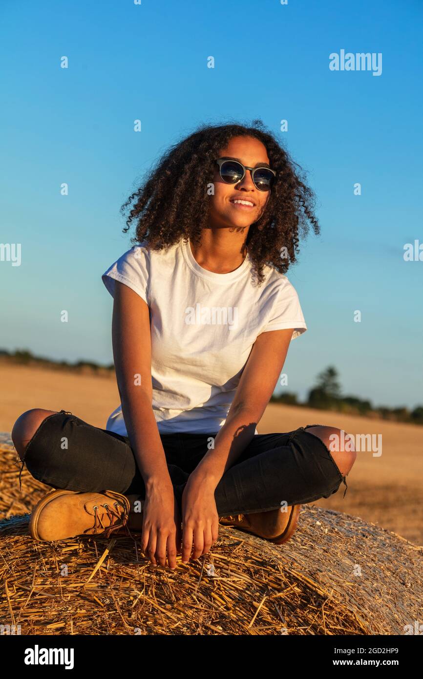 Beautiful mixed race African American female teenager young woman sitting on a hay bale in a field wearing white t-shirt and sunglasses at sunset Stock Photo