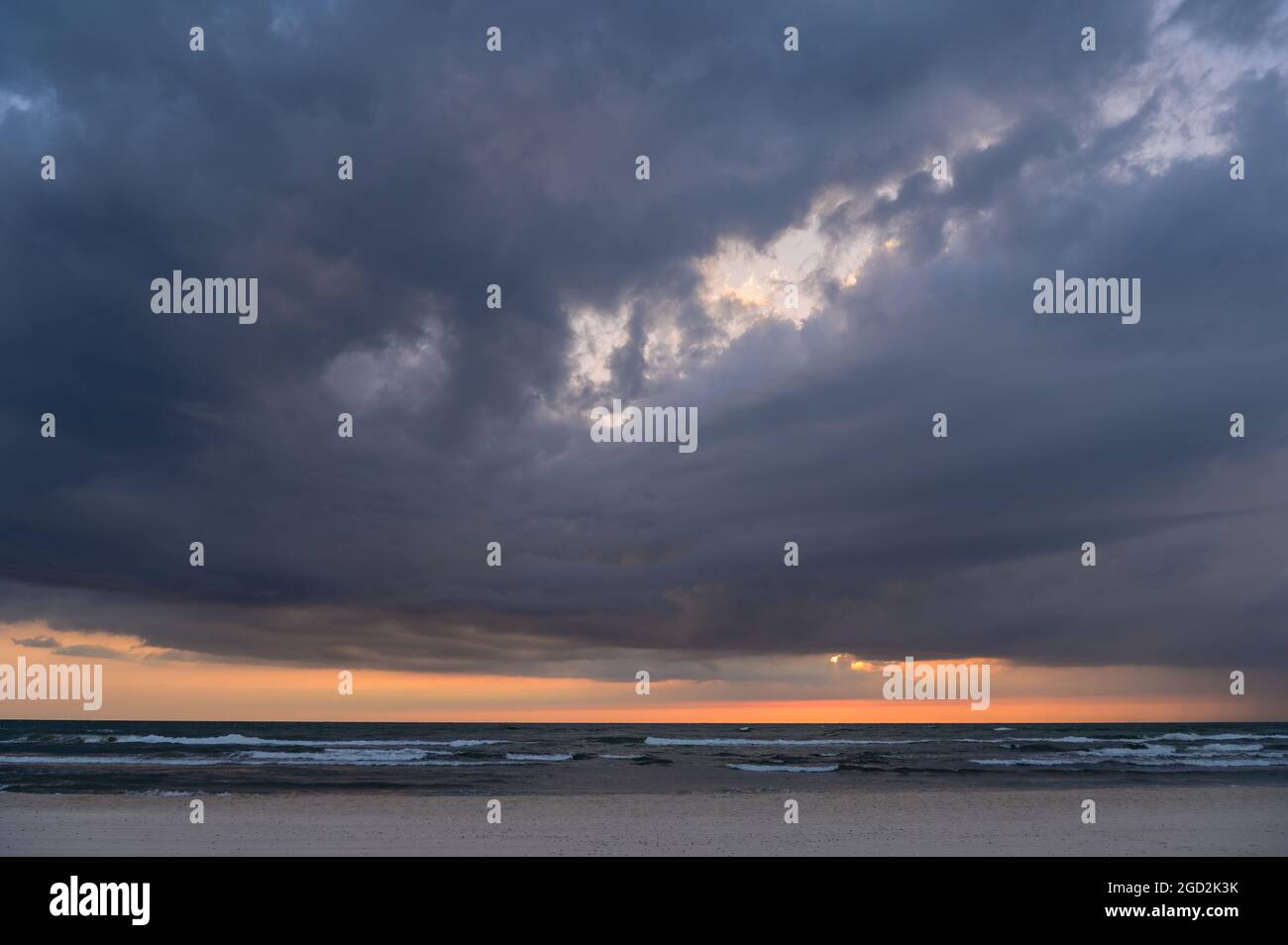 dark clouds at sunset by the sea with small waves and the sun scattered behind the clouds only leaving an orange stripe on the horizon Stock Photo