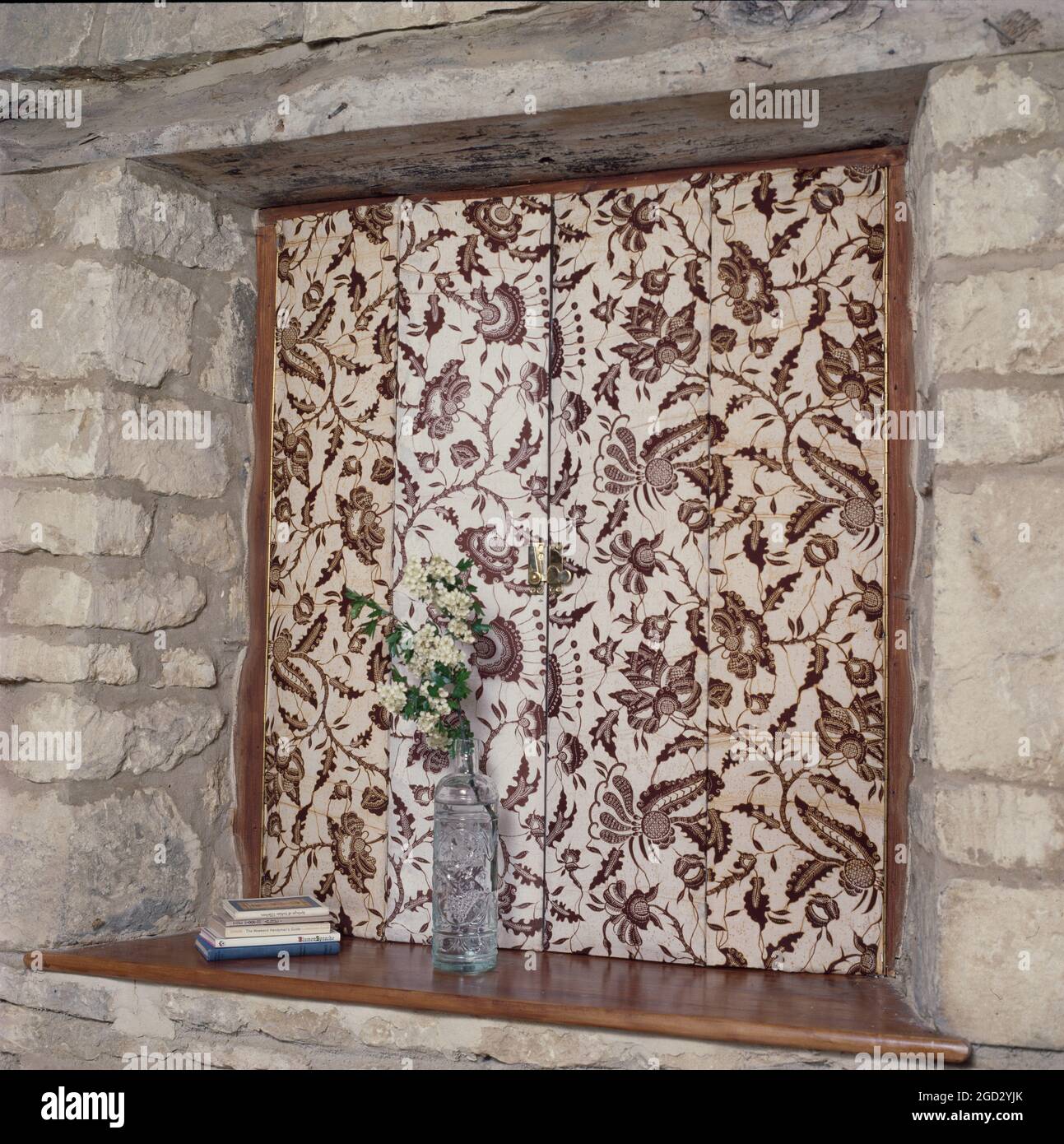 Closed floral fabric covered shutters on small window in stone wall. Stock Photo