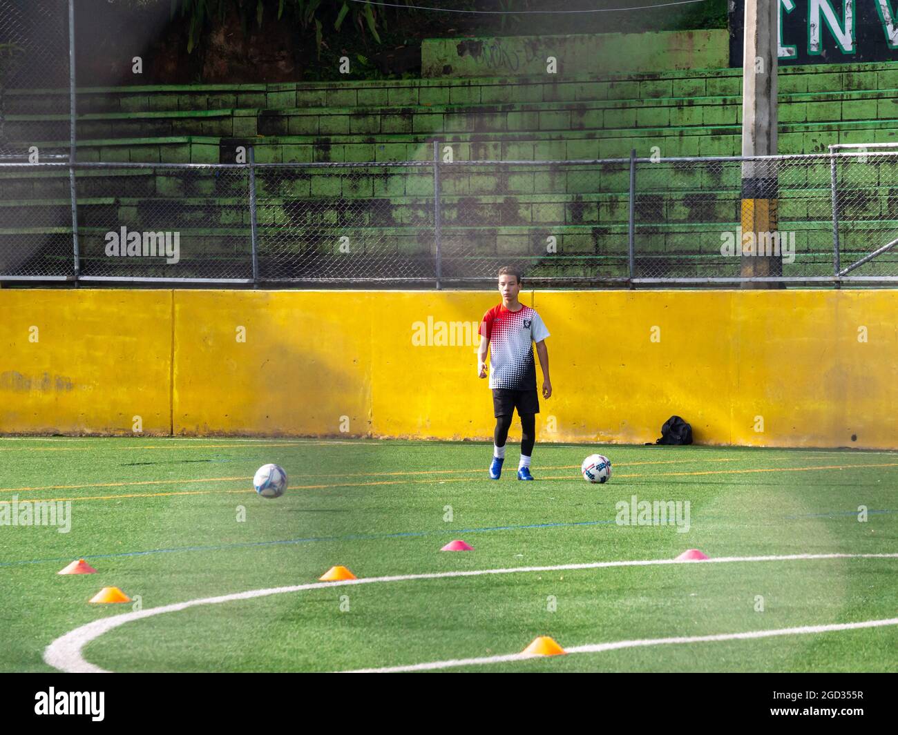 Medellin, Colombia - July 30 2021: Young Latin Teenager is Training with two Soccer Balls on a Green Synthetic Field in a Sunny Day Stock Photo