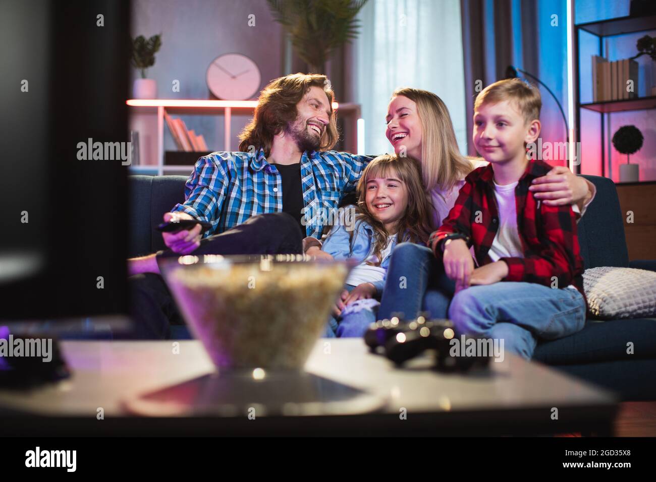 Young parents with son and daughter relaxing on comfy couch, watching TV, smiling and talking. Happy family together at home. Concept of love and unity. Stock Photo