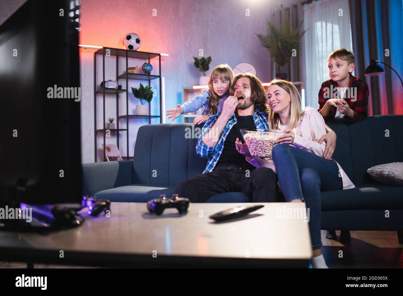 Relaxed parents with two children eating popcorn and watching TV at home. Caucasian family spending evening time together. Concept of leisure and lifestyles. Stock Photo