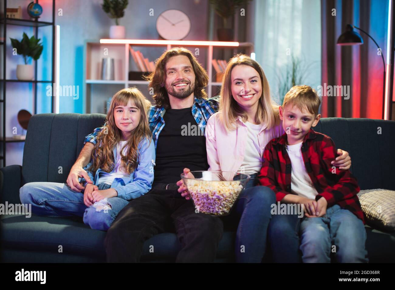 Happy caucasian family of four sitting together on couch enjoying movie with popcorn. Young parents with two kids watching TV during evening time at home. Stock Photo