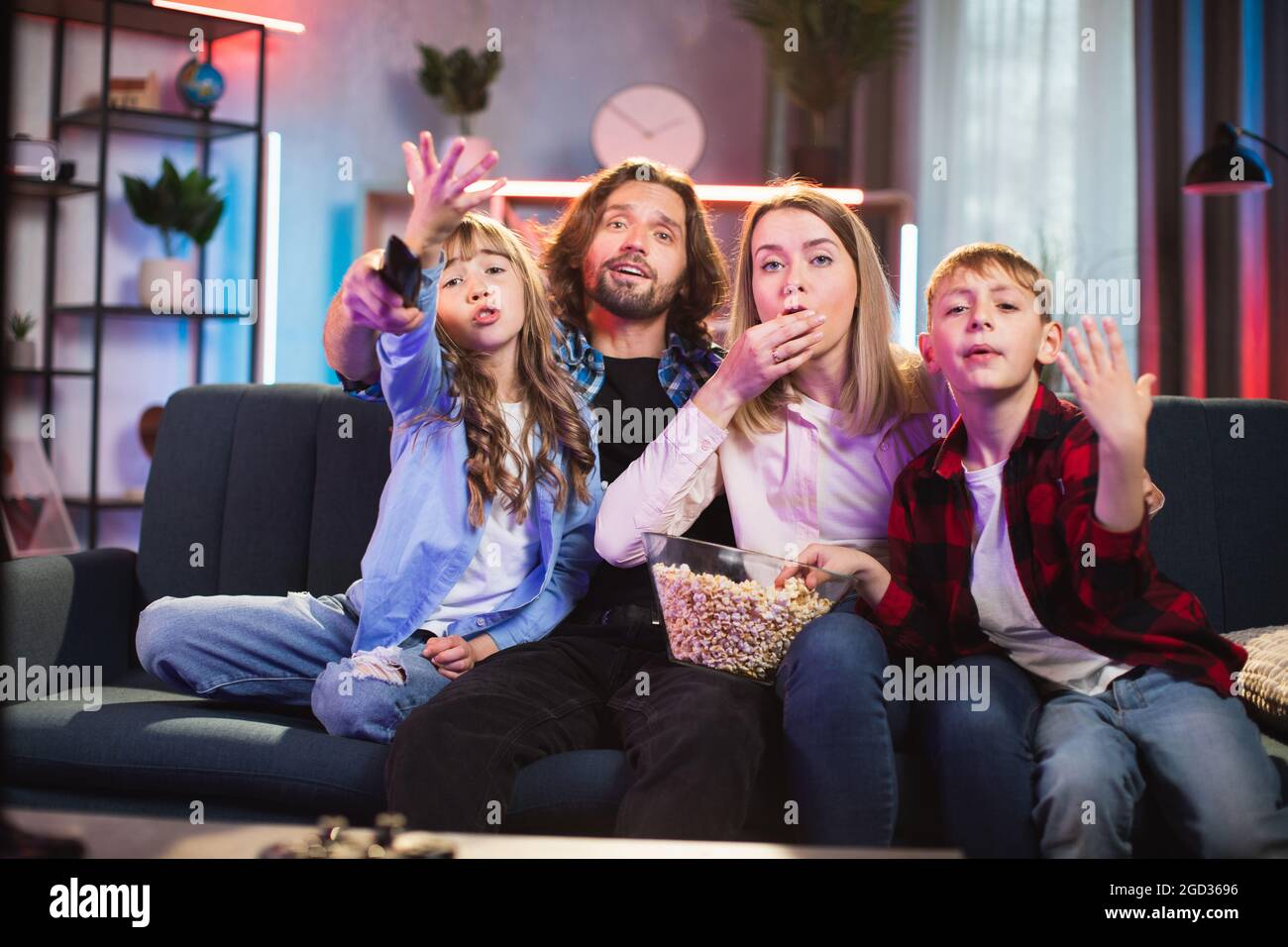 Two kids with young parents emotionally watching TV at home. Excited family in casual clothes sitting on couch and eating popcorn. Leisure time at home. Stock Photo