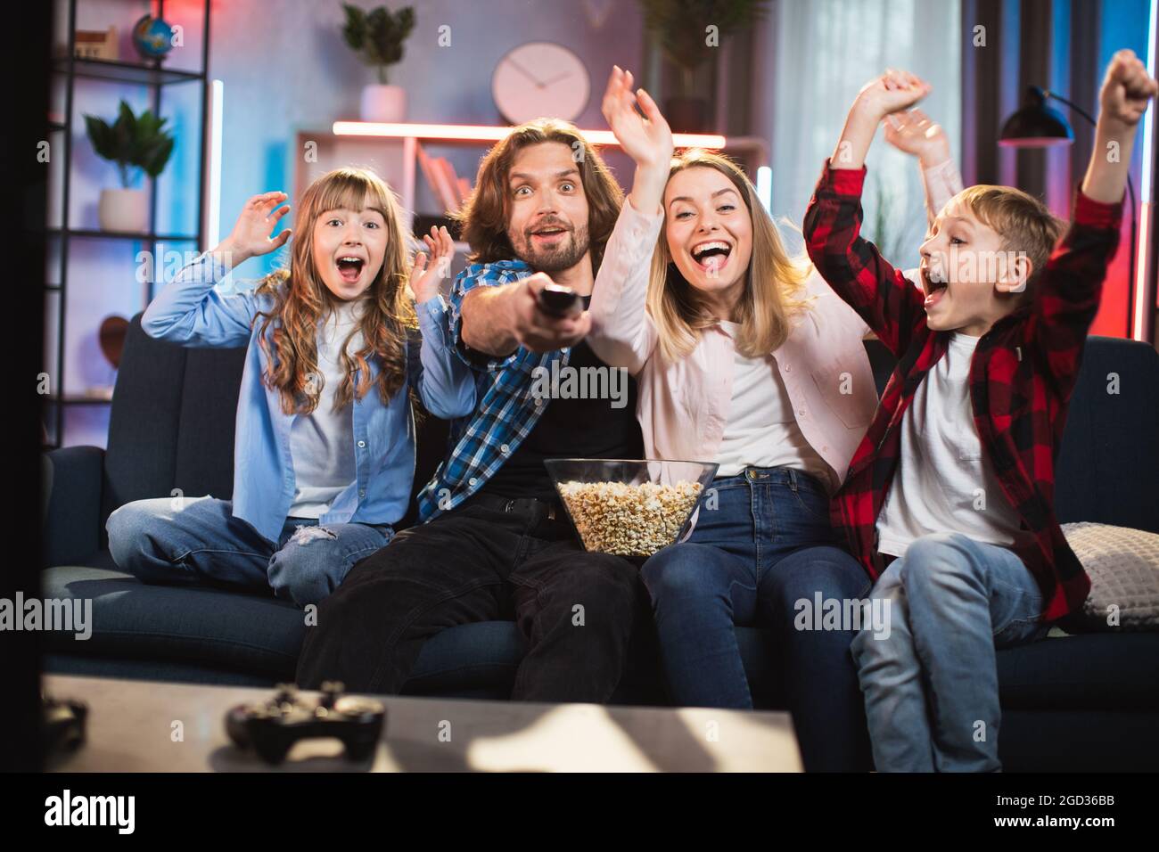 Emotional caucasian family of four dressed in casual clothes sitting on comfy couch and watching television during evening time. Human feelings and expression. Stock Photo