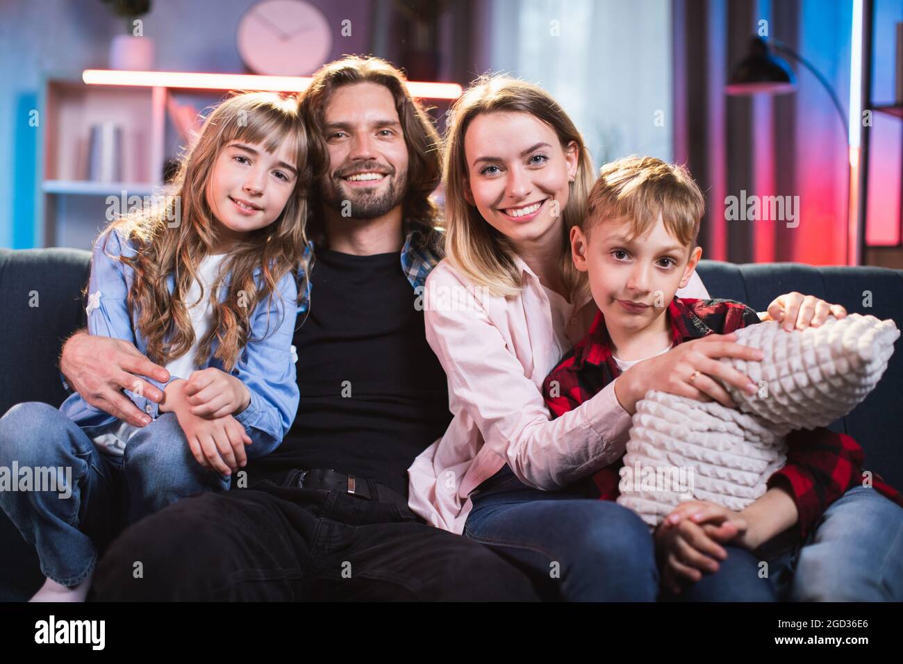 Beautiful positive family in casual clothes sitting in hugs on comfy couch and spending evening time for watching TV. Parents with kids together at home. Stock Photo