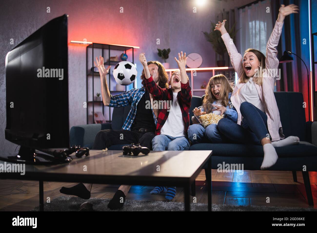 Emotional parents with little son and daughter celebrating goal of favorite team during soccer match. Satisfied family sitting on couch and watching sport competition on TV. Stock Photo