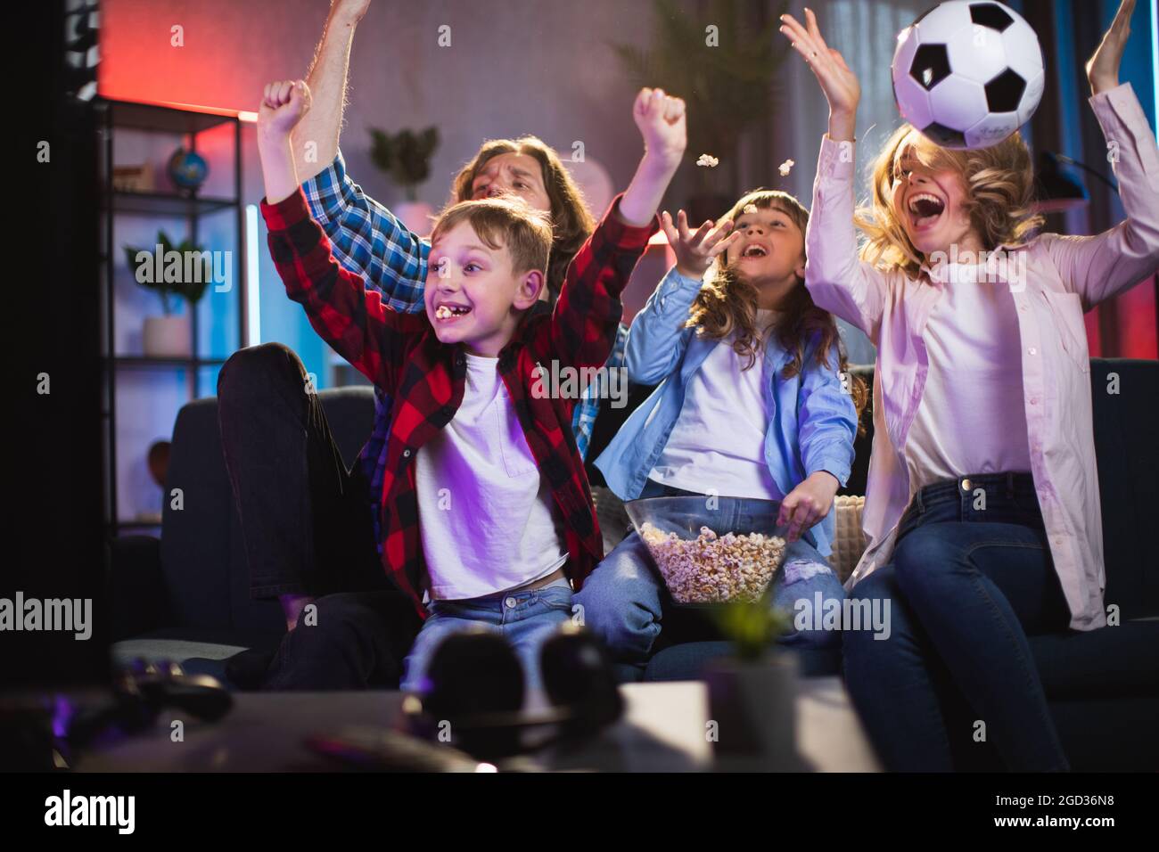 Caucasian family of four emotionally celebrating goal while watching soccer on TV. Parents with two kids sitting on couch, eating popcorn and enjoying time together. Stock Photo