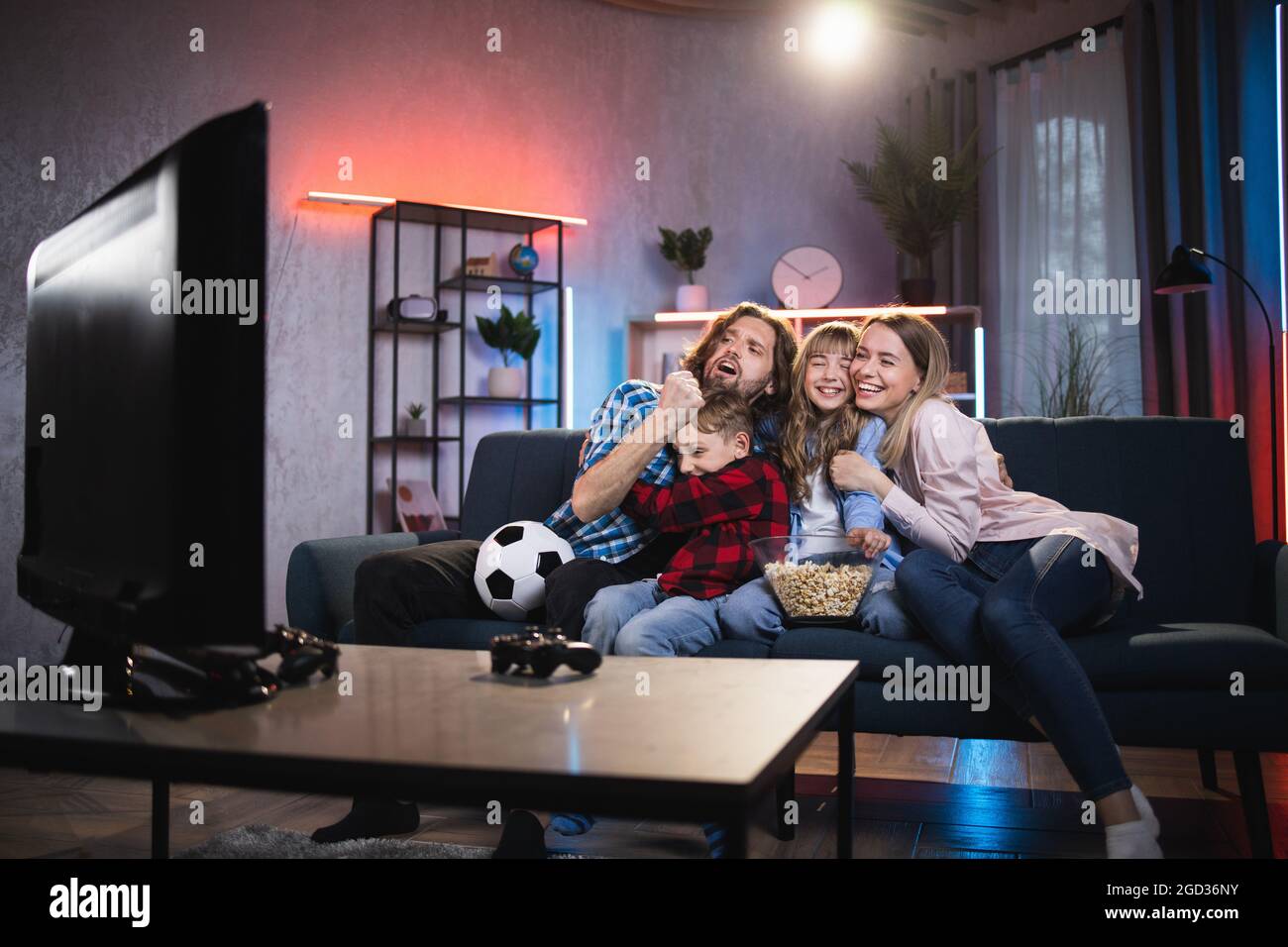 Happy young parents with son and daughter watching football game on TV. Caucasian family sitting on couch and embracing each other while celebrating goal. Stock Photo