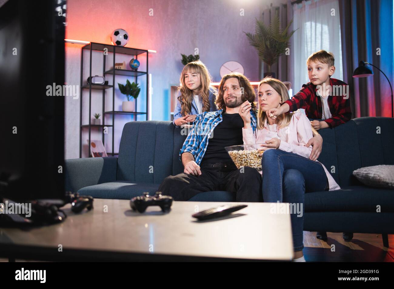 Family entertainment and unity. Relaxed happy smiling parents and their two cute kids boy and girl, watching TV-show at home in the evening and eating popcorn from glass bowl Stock Photo