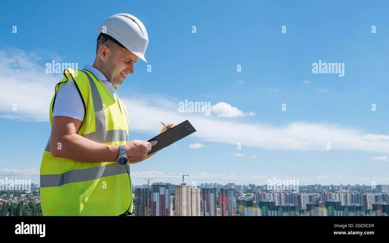 Smiling man in white helmet at construction site writes notes on clipboard Stock Photo