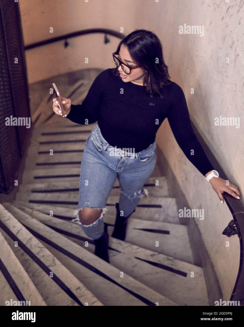 brunette girl wearing glasses climbing a spiral staircase looking at her smart phone. vertical Stock Photo