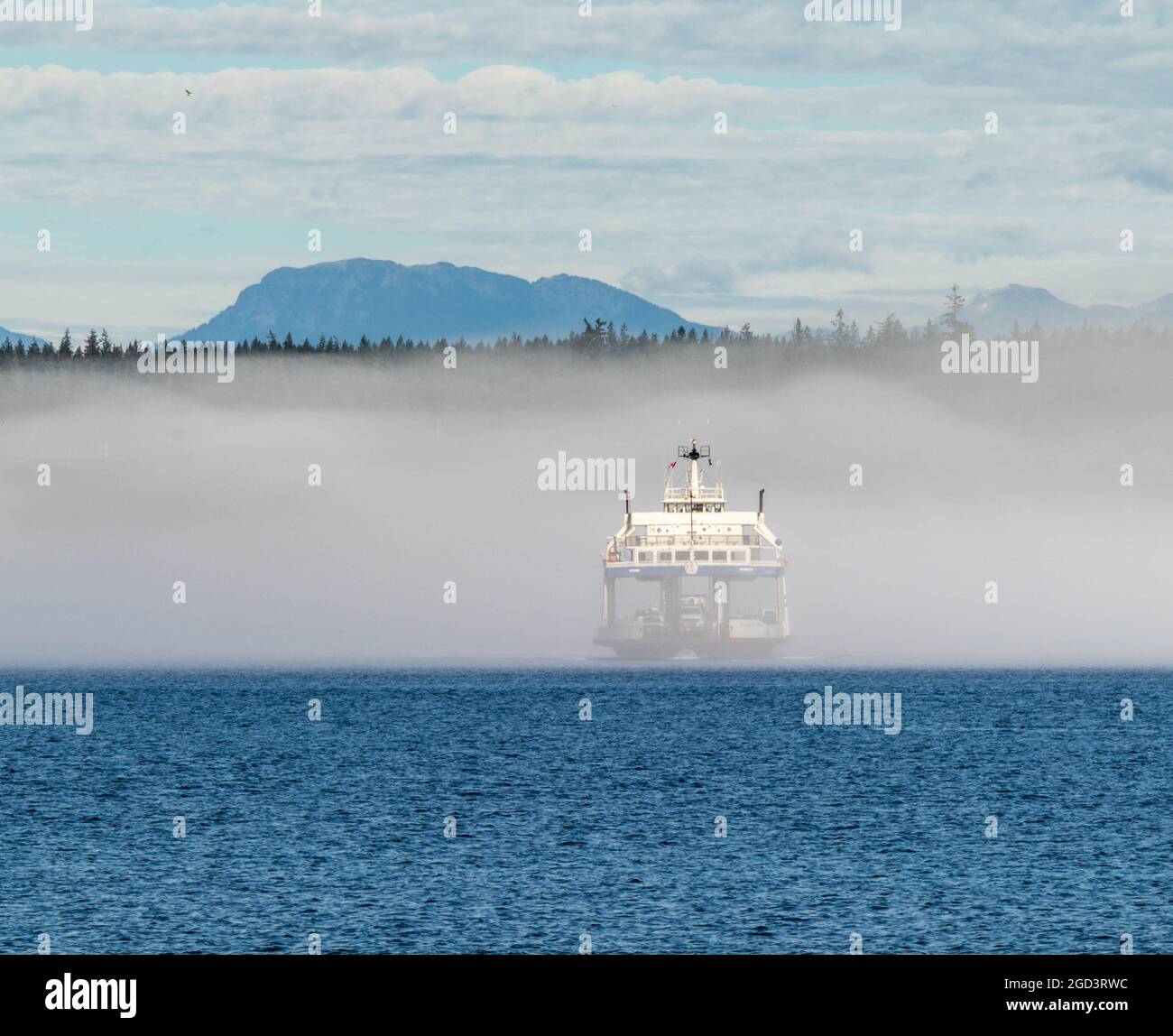 Car ferry crossing Discovery Passage in BC, Canada.  Blue water, mountain and forest in background, ferry partially obscured by fog. Stock Photo