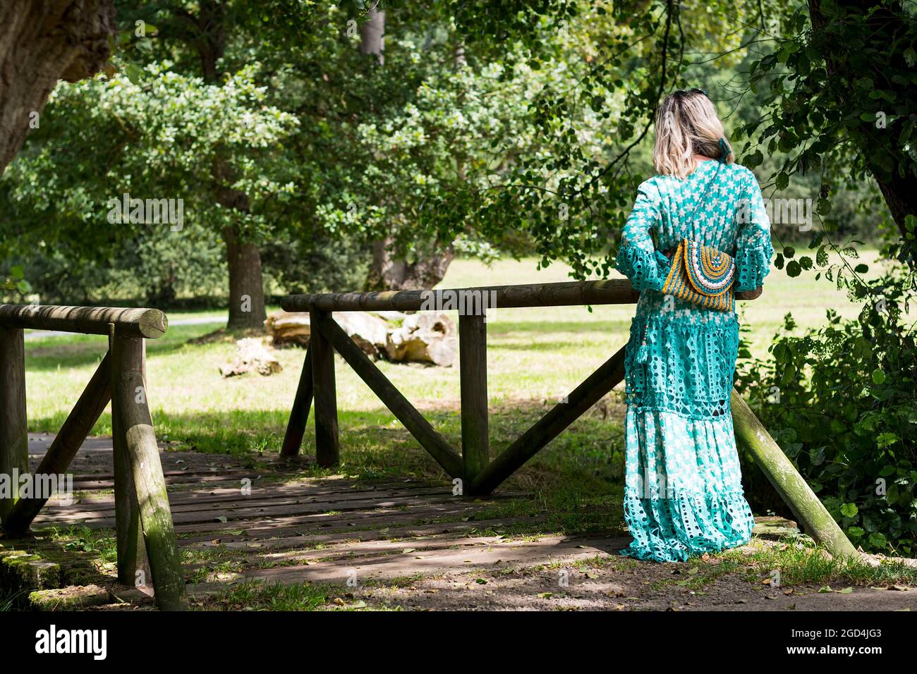 Spanish blonde woman posing with her back on a wooden bridge in a park in Asturias showing her bag and her long green dress.The photograph has a beaut Stock Photo