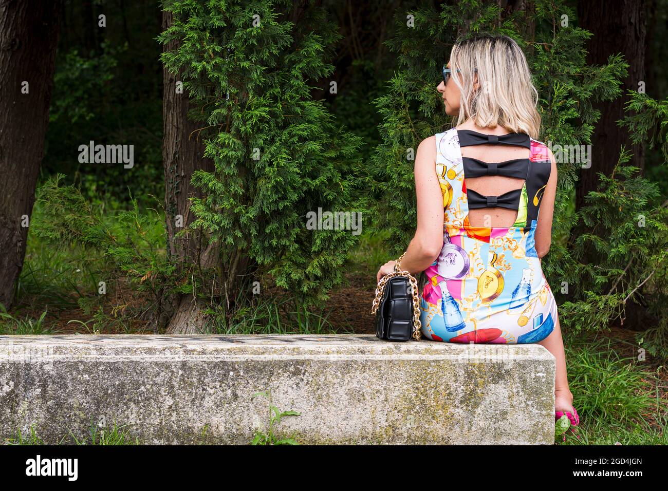 Blond woman sitting on a stone bench with her back to the camera.The model is wearing a colorful dress, pink sandals and a black bag in her hand.The p Stock Photo