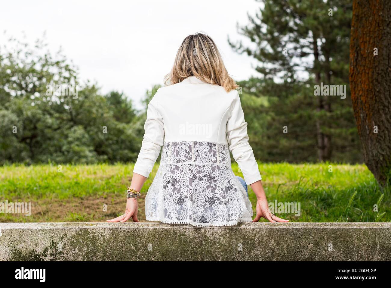 Blond haired woman sitting with her back to the camera on a stone bench in a park in Asturias, Spain.The woman is wearing jeans and a white jacket.Fas Stock Photo