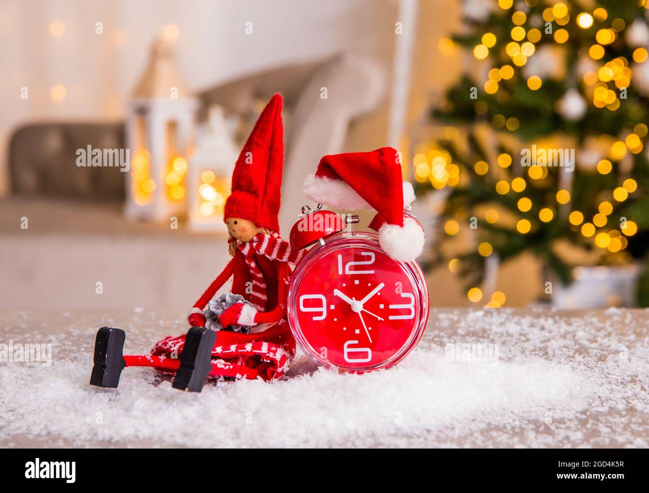 Decorative felt elf, red vintage alarm clock with small Christmas hat on artificial snow, blurred Christmas tree with gifts and white illuminated. Stock Photo