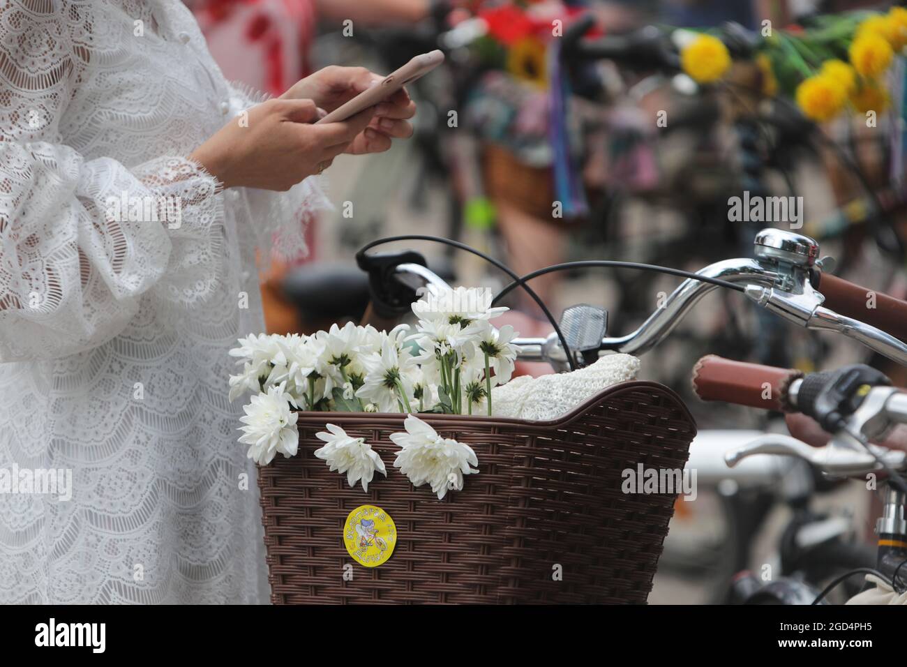 KYIV, UKRAINE - AUGUST 7, 2021 - A woman in a white lace dress browses her smartphone next to a basket with white flowers attached to a bicycle as see Stock Photo