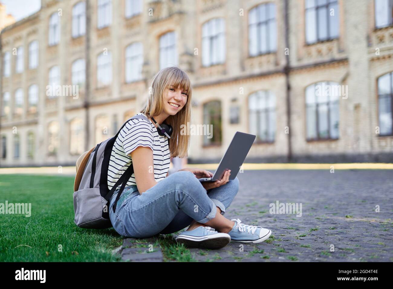 Young cute caucasian blonde female student at university campus sitting on green grass with backpack and laptop browsing internet happy emotion looking straight at camera. High quality photo Stock Photo