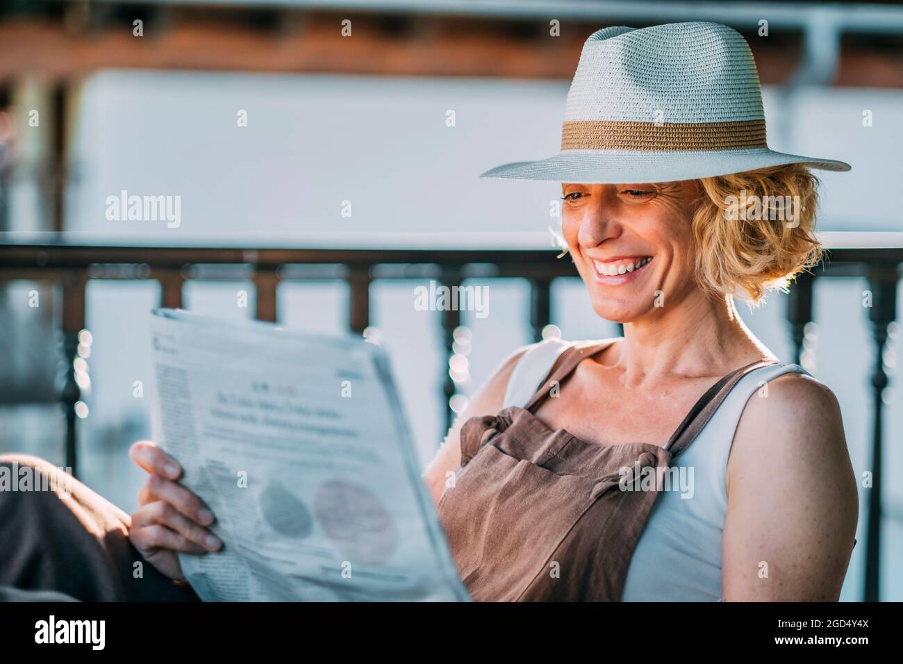 Mature caucasian blonde woman in her 50s with a hat and reading a newspaper sitting in a rocking chair in a relaxed pose on a porch. Lifestyle concept. Stock Photo