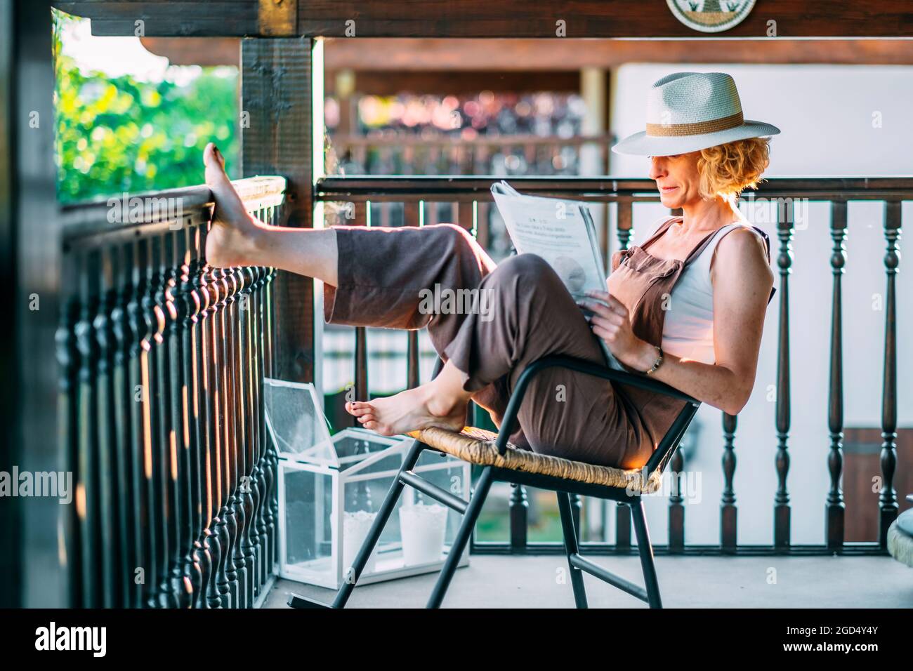 Mature caucasian blonde woman in her 50s with a hat and reading a newspaper sitting in a rocking chair in a relaxed pose on a porch. Lifestyle concept. Stock Photo