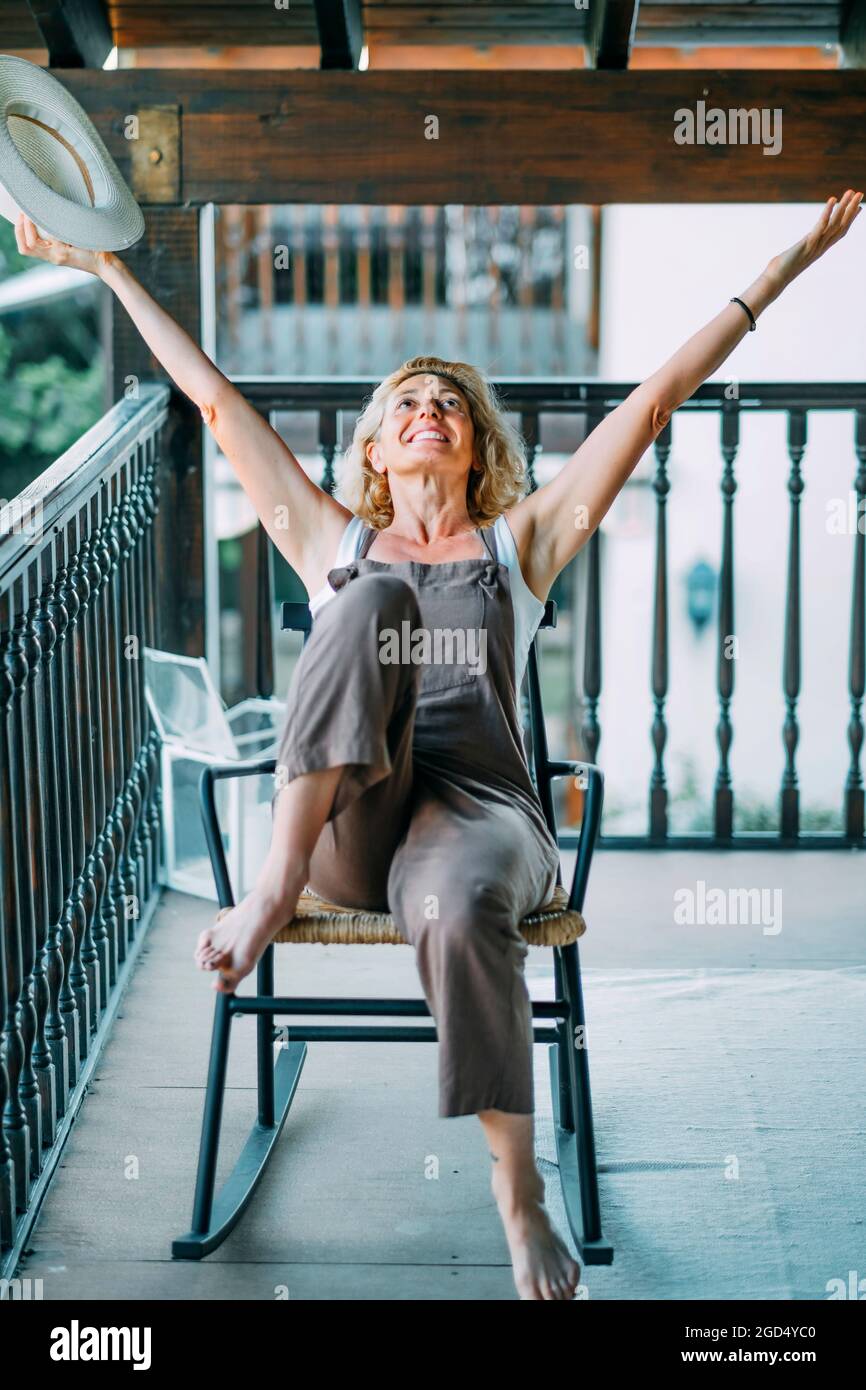 Mature caucasian blonde woman in her 50s with hat and sitting in a relaxed  pose in a rocking chair on the porch of a country house. Lifestyle concept  Stock Photo - Alamy