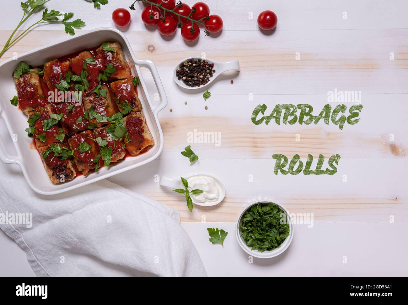 Square baking pan with cabbage rolls in tomato sauce, cherry tomatoes, chopped fresh parsley, sour cream, linen napkin. Flat lay with. White and beige Stock Photo