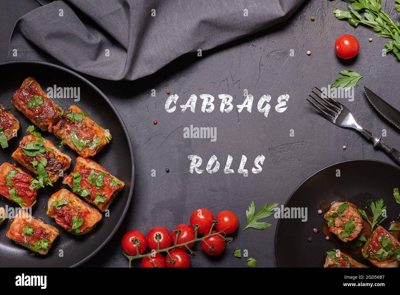 Flat lay with cabbage rolls with tomato sauce and chopped parsley on frying pan and on black plate and also cherry tomatoes, metal fork and knife, lin Stock Photo