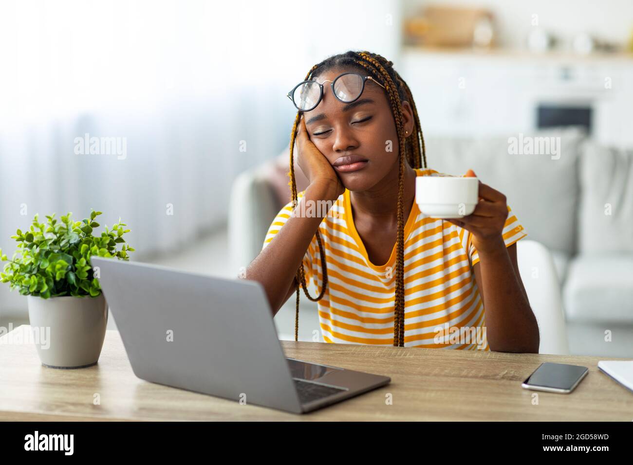 Exhausted millennial black woman freelancer sitting in front of laptop with cup of coffee, feeling sleepy and tired or hangover, suffering from burnou Stock Photo