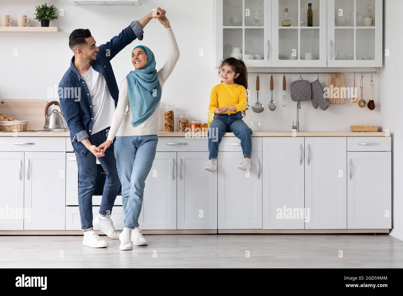 Portrait Of Happy Arab Family Of Three Having Fun In Kitchen Interior, Cute Smiling Little Middle Eastern Girl Sitting On Table And Watching Her Paren Stock Photo