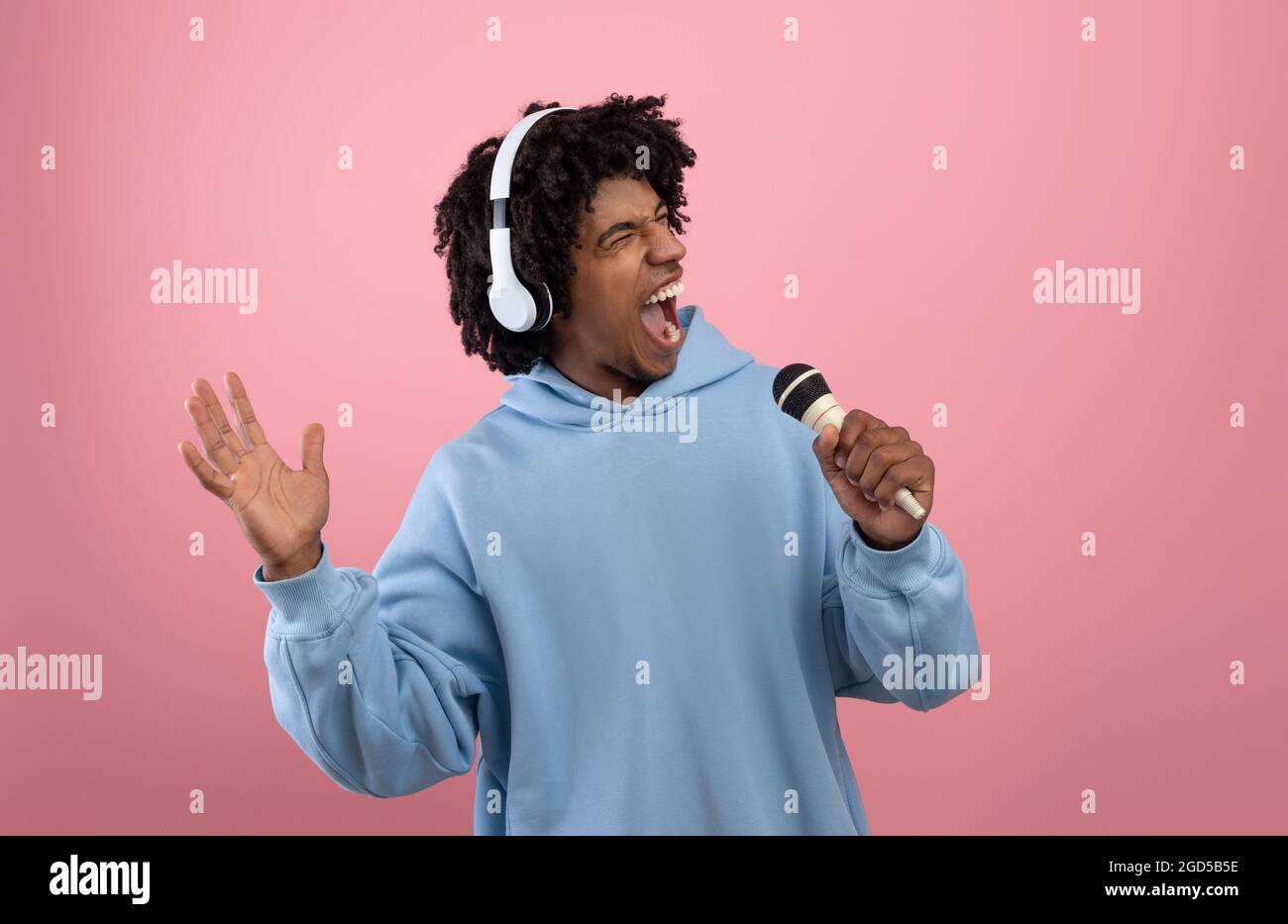 Creative Afro teen in headphones listening to music, singing song into microphone on pink studio background. Black adolescent in headset pretending to Stock Photo