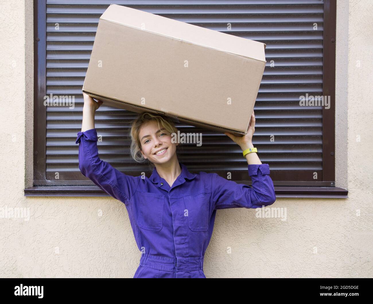 young smiling woman in a purple uniform holds a cardboard box on her head. Delivery, surprise, beautiful courier. Business, mail, gift. Part-time jobs Stock Photo