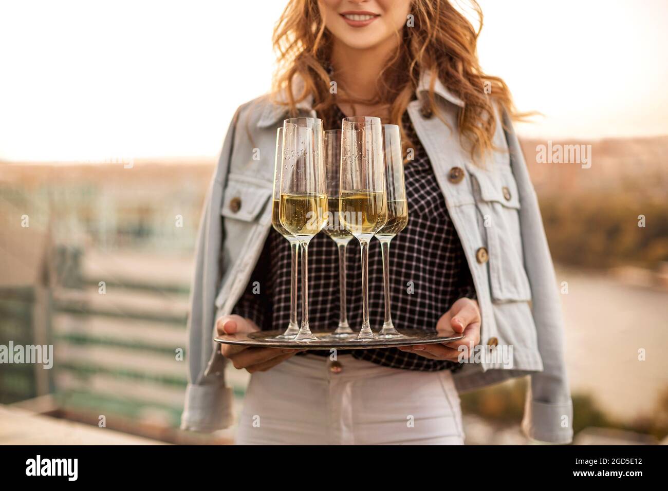 Young blonde smiling woman holding tray with champagne sparkling wine in flute glasses, standing outdoors over blurred countryside background, celebra Stock Photo