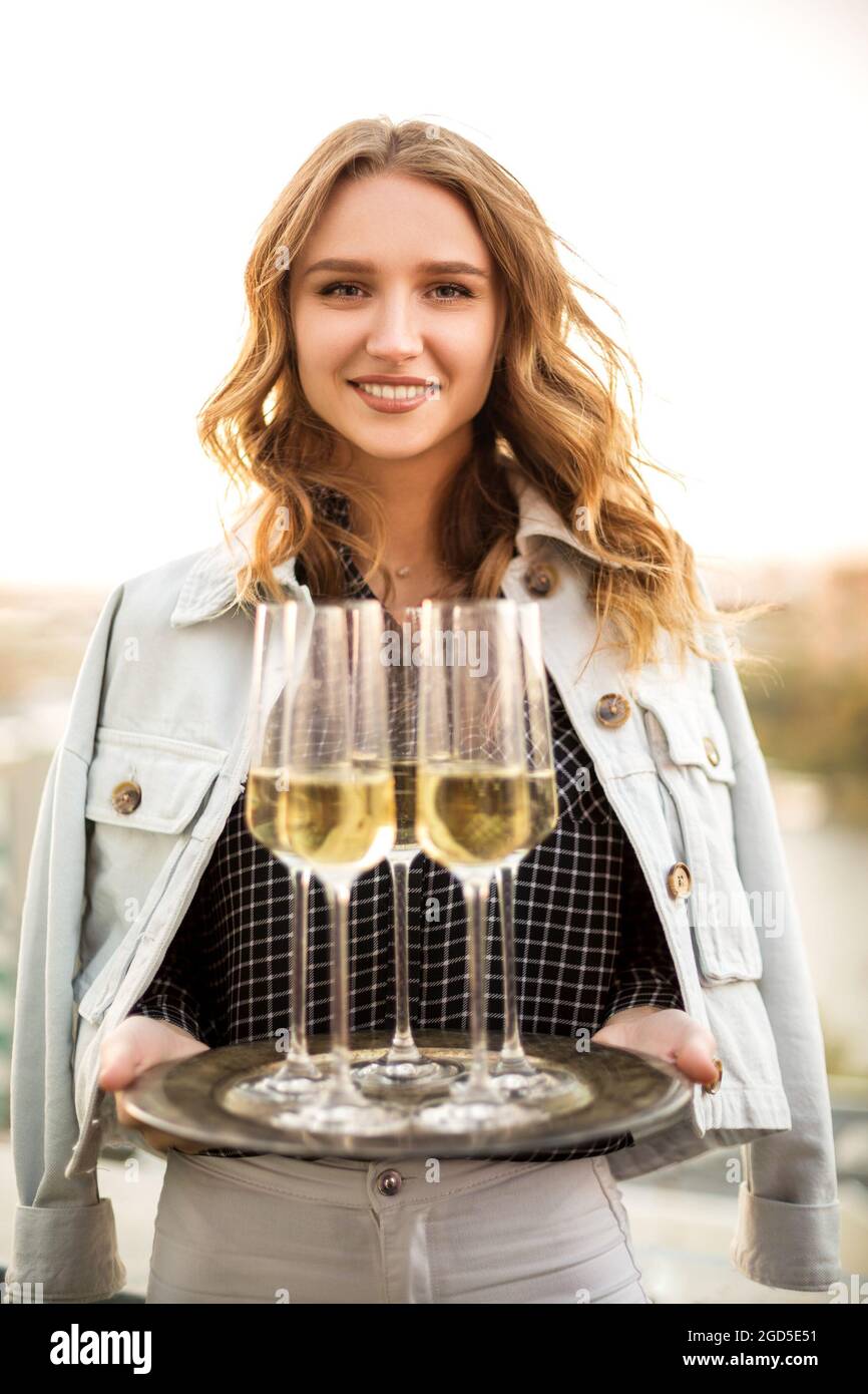 Young blonde smiling woman holding tray with champagne sparkling wine in flute glasses, standing outdoors over blurred countryside background, celebra Stock Photo
