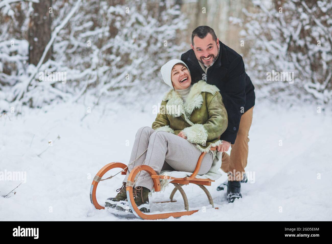 Happy playful mature family couple sledding in winter park, laughing and having fun together, positive middle-aged woman sitting on sled while her hus Stock Photo