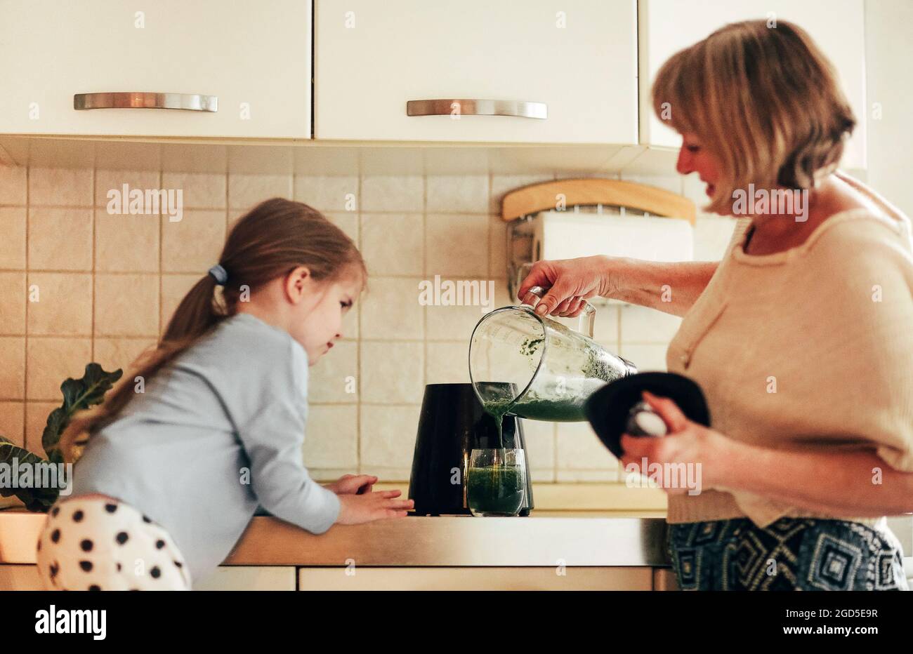 Loving grandmother cooking together with granddaughter at home, using blender to mix vegetables, little girl learning how to use appliances with grand Stock Photo