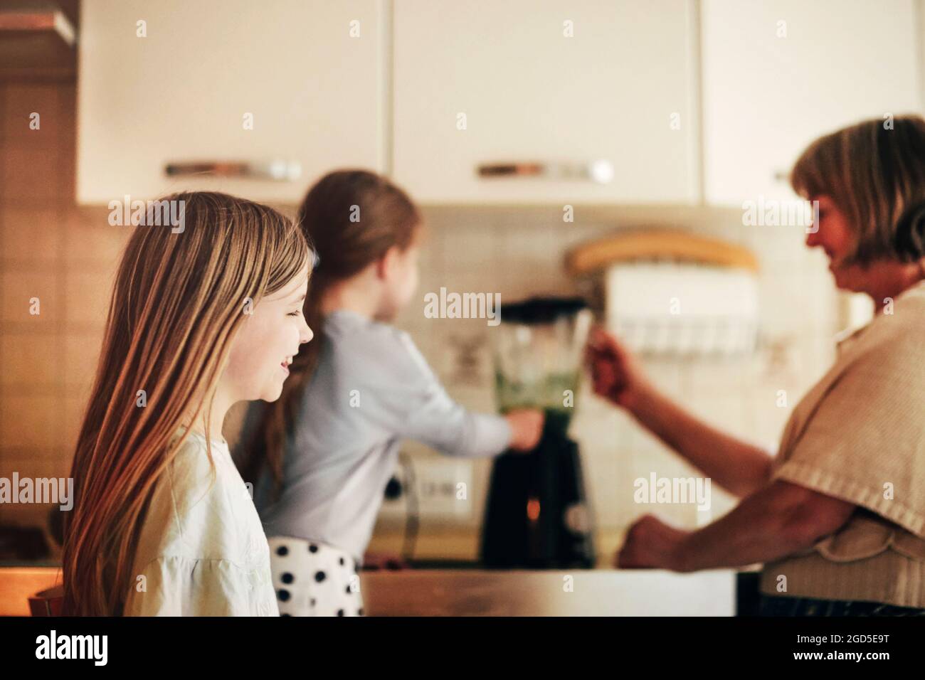 Loving grandmother cooking together with granddaughters at home, using blender to mix vegetables, little girl learning how to use appliances with gran Stock Photo