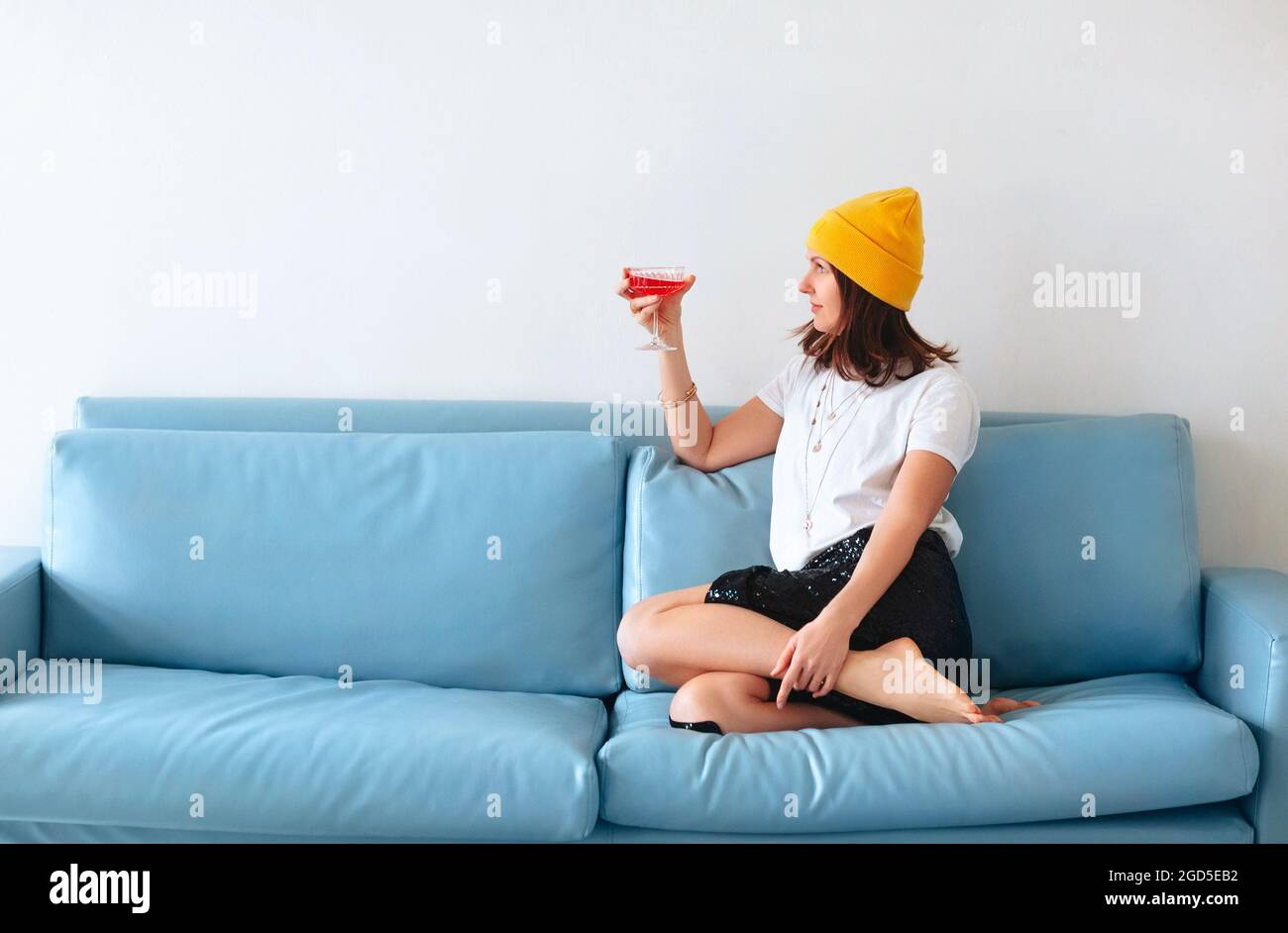 Cropped shot of young smiling woman holding cocktail glass in hand with red alcohol drink sitting on sofa at home, wearing fashionable clothes and acc Stock Photo