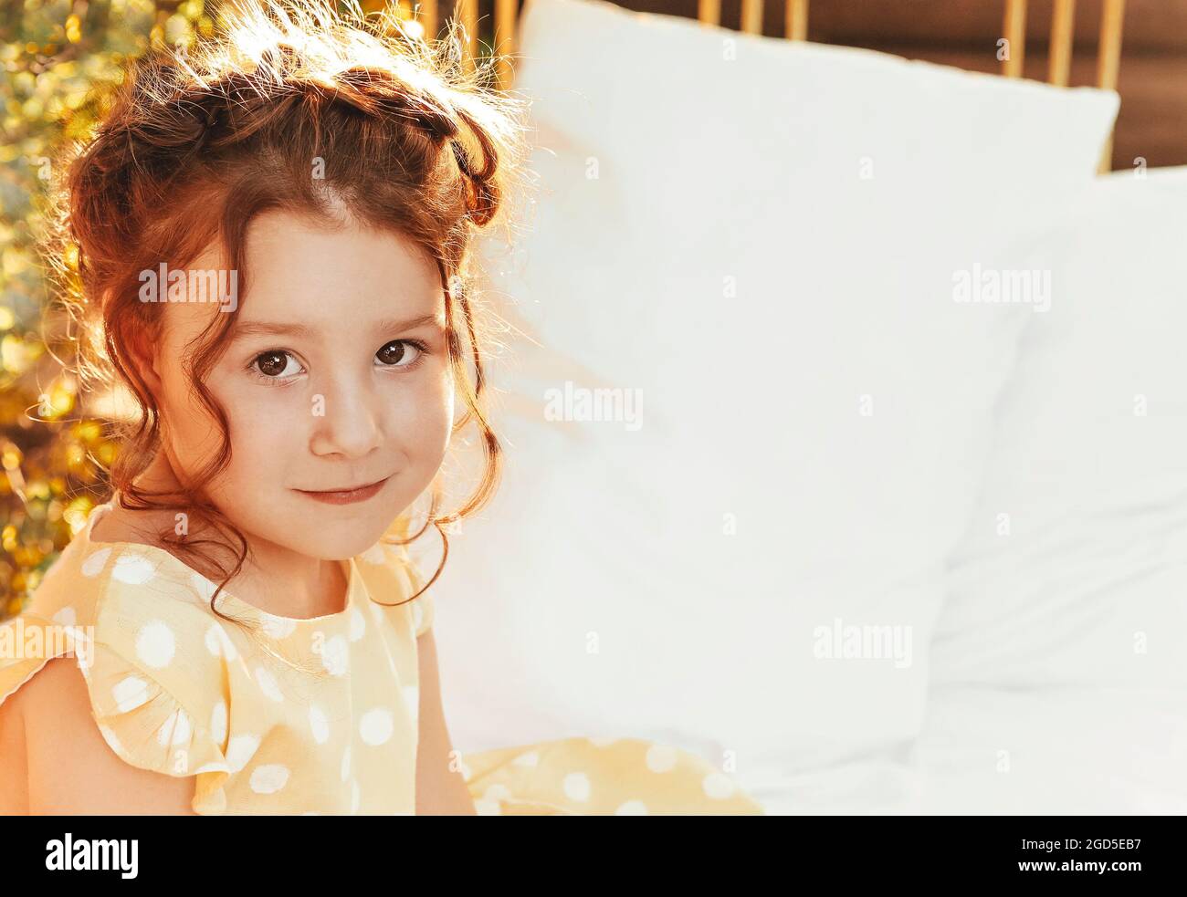 Close up portrait of cute little adorable redhead girl with curly hair with braid wearing yellow dotted dress looking at camera while playing in natur Stock Photo