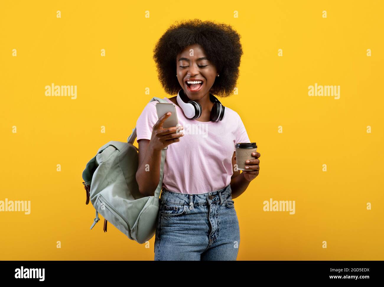 Online education. Happy black lady with backpack, headphones, cellphone and takeaway coffee standing over yellow studio background. Young female stude Stock Photo