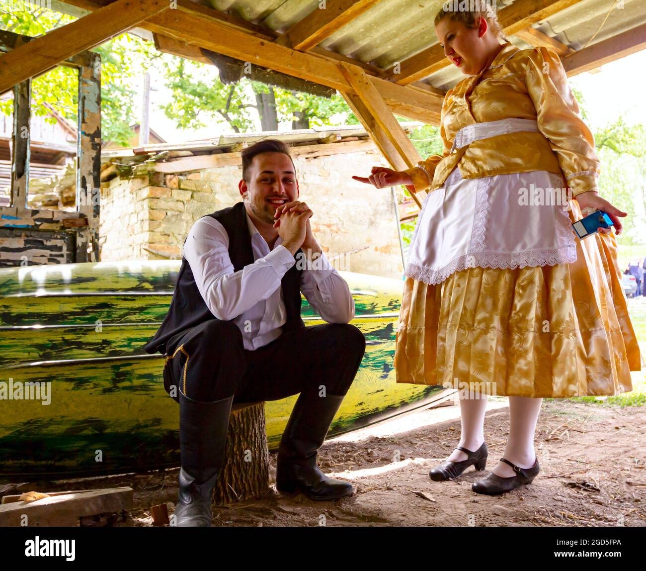 Ivanovo, Vojvodina, Serbia - April 17, 2016: Girl make jokes with young man who is sitting on wooden stump wearing a traditional folk costume in ancie Stock Photo