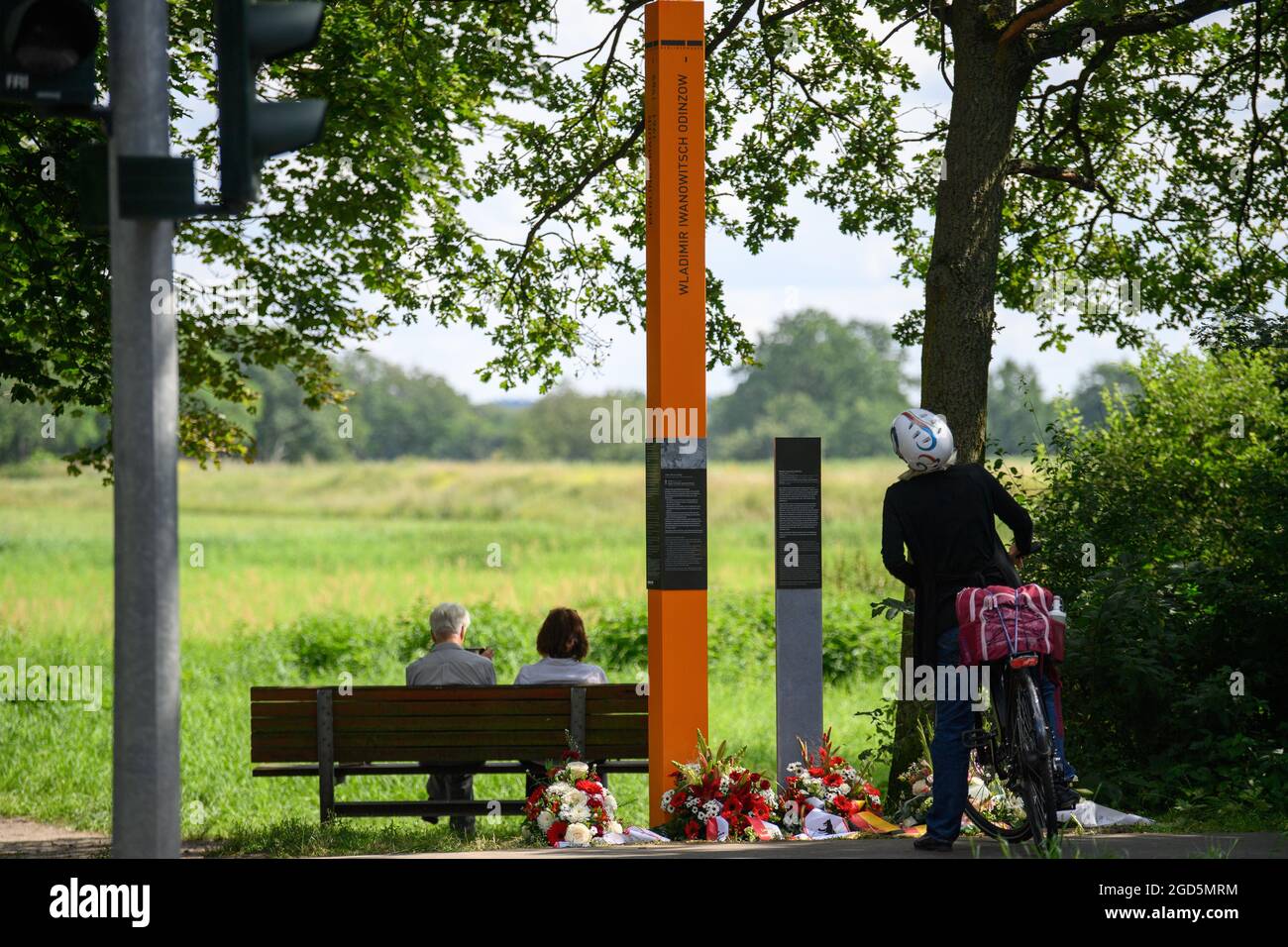 11 August 2021, Brandenburg, Dallgow-Döberitz/Ot Seeburg: A man and a woman sit on a bench behind the memorial stele for Vladimir Ivanovich Odintsov. The stele had been inaugurated by Brandenburg's state parliament president Liedtke. Odintsov was a Soviet soldier stationed in the artillery regiment of the Elstal garrison who had been accidentally shot by GDR People's Police officers. The policemen had mistaken him for a soldier who had been put on the wanted list. Photo: Soeren Stache/dpa-Zentralbild/dpa Stock Photo