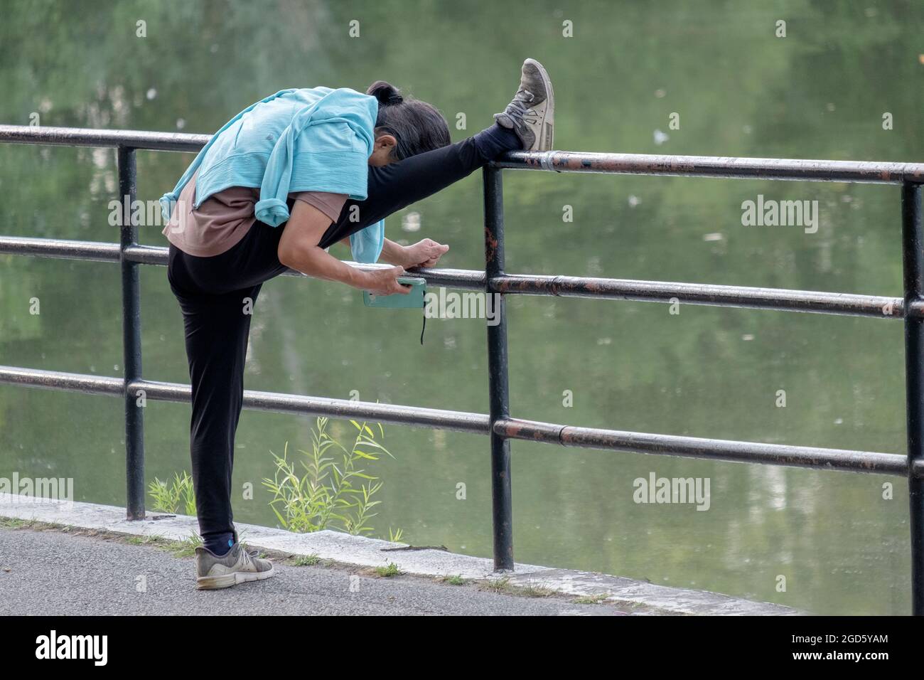 MULTITASKING. A woman stretches her hamstring and reads her cell phone at the same time. In Kissena Park in Flushing, Queens, New York City. Stock Photo
